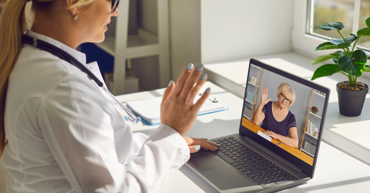 A healthcare professional conducts a virtual consultation using a healthcare communication platform. The female doctor, visible on the left, is speaking to an elderly female patient who appears on the laptop screen, smiling and waving. The setting is a bright, modern office with a plant and documents, highlighting the convenience and personal connection enabled by telehealth technologies.