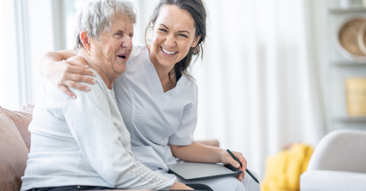 A compassionate healthtech scene where a young female healthcare worker, wearing a white uniform, smiles joyfully while embracing an elderly woman in a cozy, well-lit living room. The elderly woman, appearing pleased and comfortable, listens intently to the healthcare worker who is holding a digital tablet, ready to assist with health management.