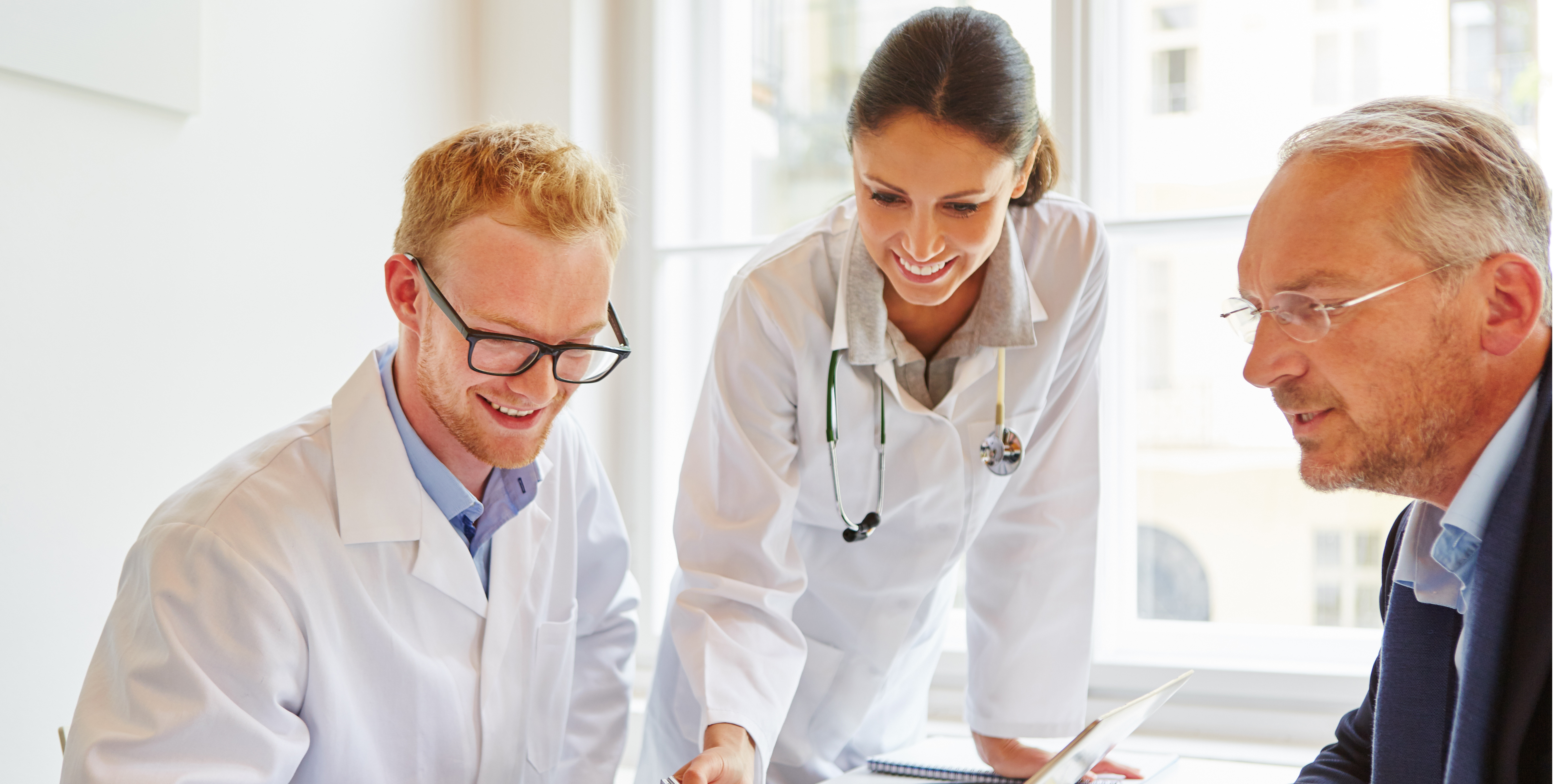Three healthcare professionals, two wearing white lab coats and one in business attire, collaboratively discuss over a digital tablet in a bright office setting. They represent a dynamic team utilizing modern communication tools in a healthcare environment