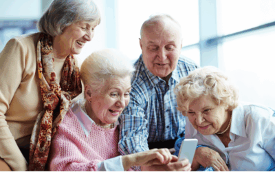 Four joyful seniors gather around a smartphone, with two women seated and looking intently at the screen while the other two, a woman and a man, stand behind them smiling.