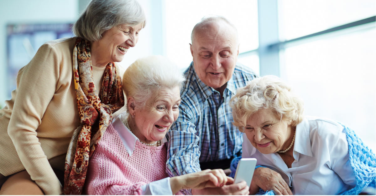 Four joyful seniors gather around a smartphone, with two women seated and looking intently at the screen while the other two, a woman and a man, stand behind them smiling.